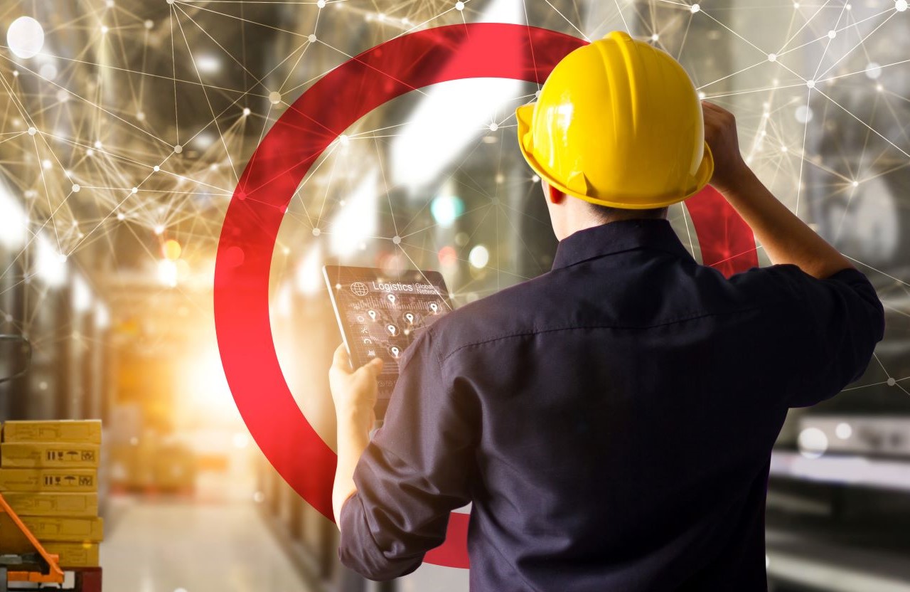 Man with yellow hard holding tablet looking ahead in warehouse setting