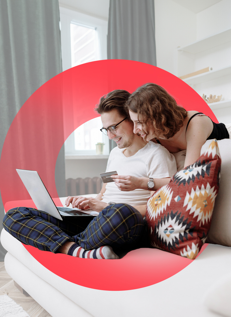 Casual smiling couple making online purchase. Man seated on couch with laptop and woman leaning over while holding credit card.