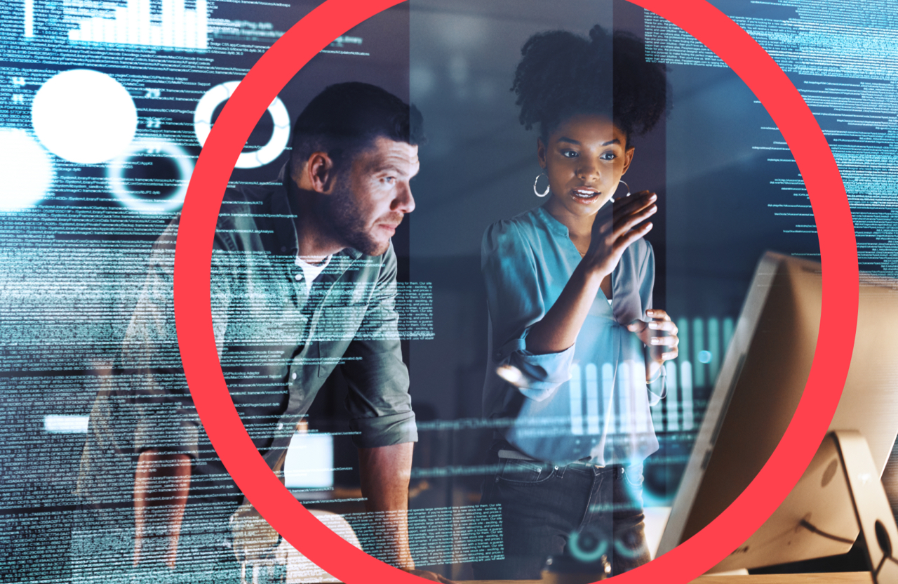 Woman standing with right arm extended towards computer monitor and standing next to man who is looking at and leaning towards computer monitor to review data projected against backdrop.