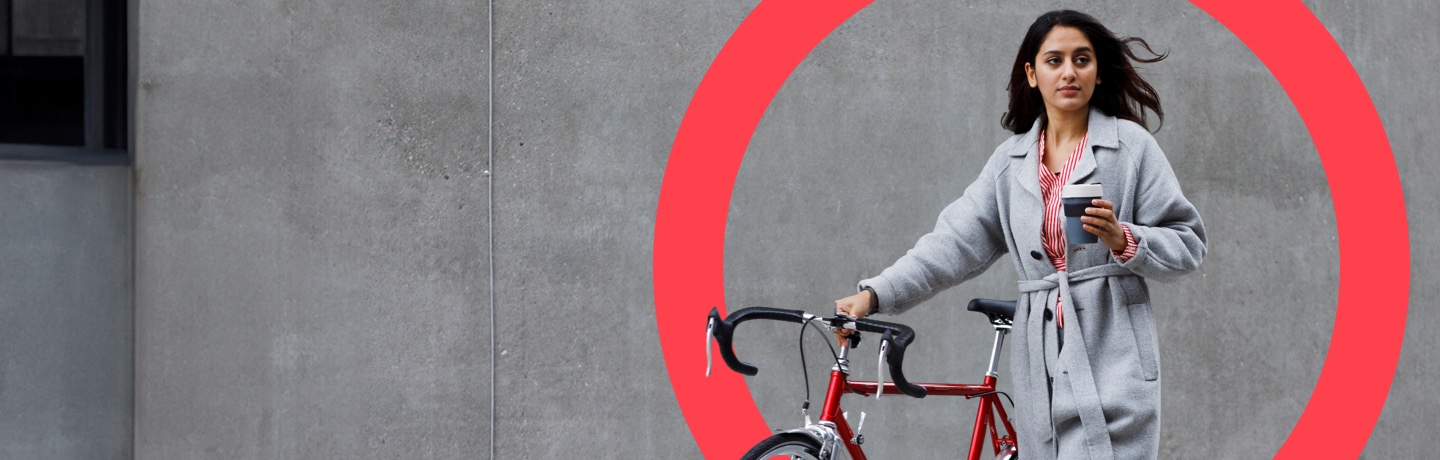 A woman in a grey coat with a bicycle, holding a cup of coffee in front of a large red circle.