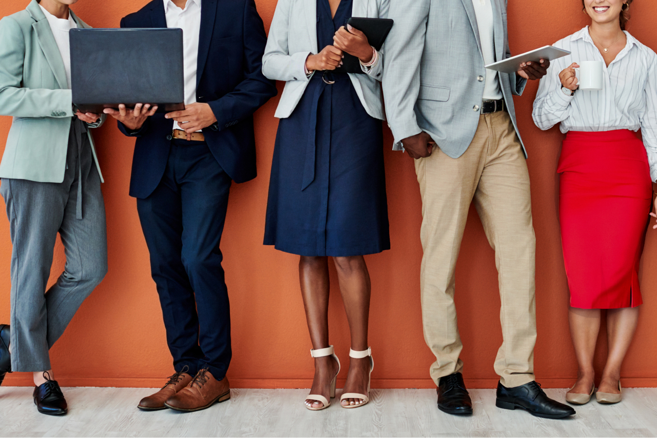 A diverse group of five people wearing business attire stand in front of an orange wall. They hold various items including laptops, tablets and coffee.