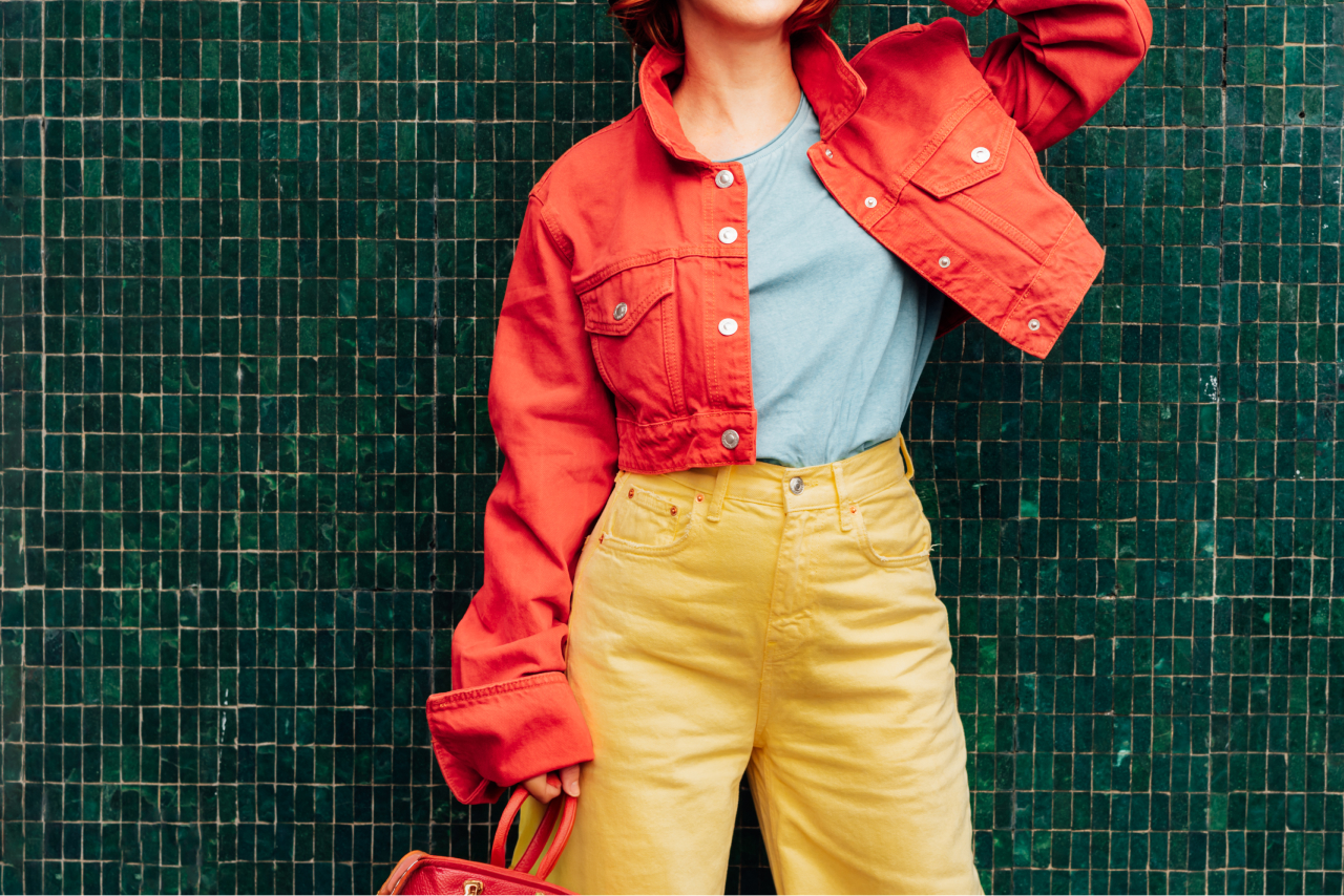 A woman wearing yellow pants and a red jacket stands with her purse in front of a green tiled wall. 