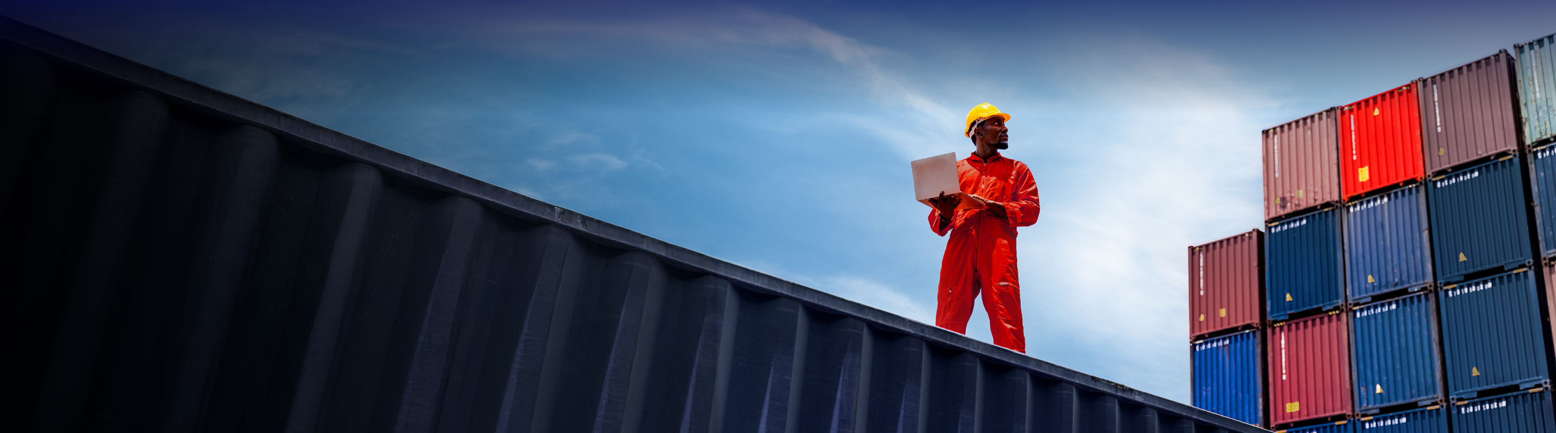 A man in overalls with a hard hat and a laptop standing on a shipping container against a blue sky